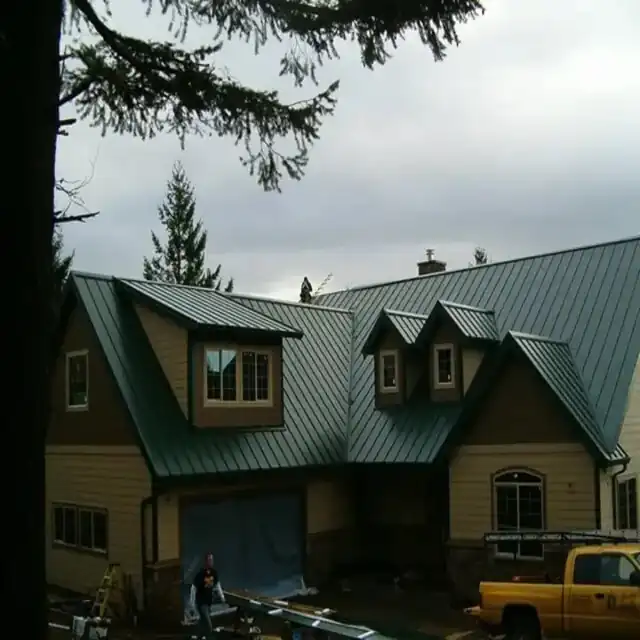 Green standing-seam roof on a dormered house with a yellow Gregg Roofing truck at the right